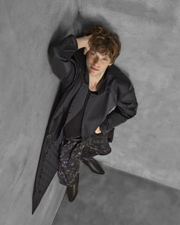 Eye-level shot of a light-skinned male model with curly hair in a black jacket and printed pants leaning against a grey corner.