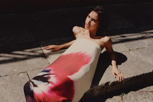 Adult woman in a strapless dress with red and black patterns lies on a stone surface outdoors in full sunlight, eyes open.
