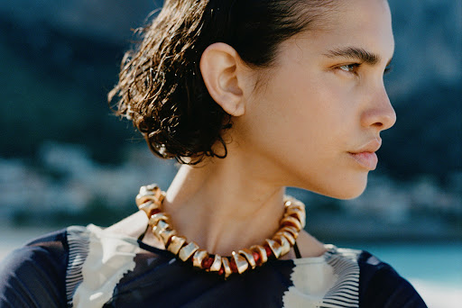 Profile shot of a woman with short, dark, wet hair, wearing a gold choker necklace, against a background of blue water and distant mountains.