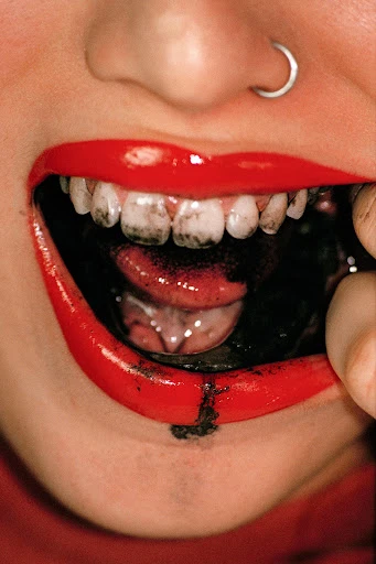 Close-up of a person's mouth with red lipstick, visible teeth, tongue, and a black substance inside the mouth and on the lips.