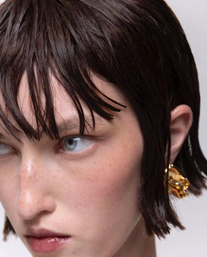 Close-up shot of an adult woman with dark, wet hair, light eyes, wearing a gold earring, beauty shot, neutral expression, plain background.