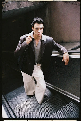 Stylish man on an escalator: He wears a brown leather jacket, grey collared shirt, and cream-colored pants, carrying a black bag over his shoulder.