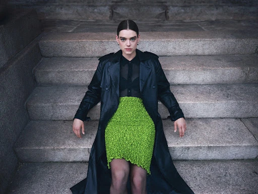 A woman in a black coat and bright green skirt sits on grey stone steps in an outdoor setting, captured in a medium shot.