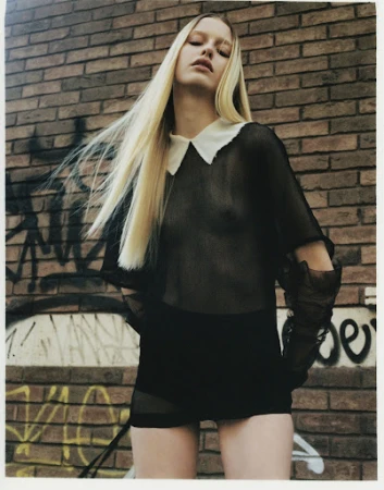 Model with long blonde hair wearing a sheer black top with a white collar against a brick wall with graffiti