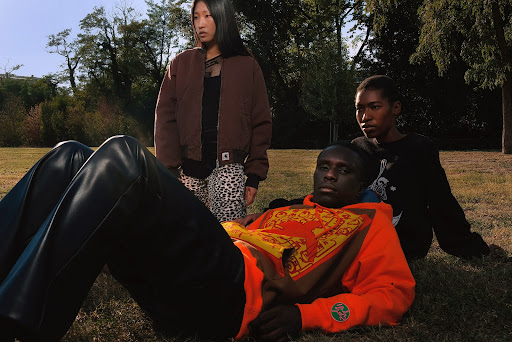Three models in outdoor setting, one lying down wearing an orange sweatshirt, with black pants. The other two standing in park.