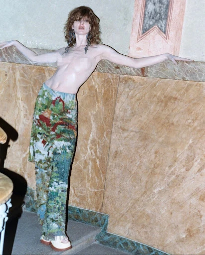 A woman poses topless in patterned pants on a marble staircase. The indoor fashion shot features natural light and earthy tones.