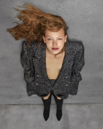 Red-haired woman wearing a dark tweed coat and tall black boots lying against a textured grey background