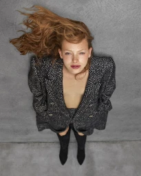 Red-haired woman wearing a dark tweed coat and tall black boots lying against a textured grey background