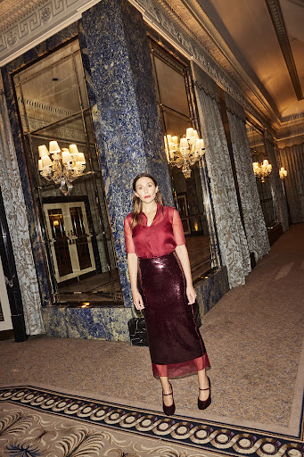 A brunette woman poses in a red blouse and shiny skirt in a grand hall with ornate pillars, chandeliers and mirrors.