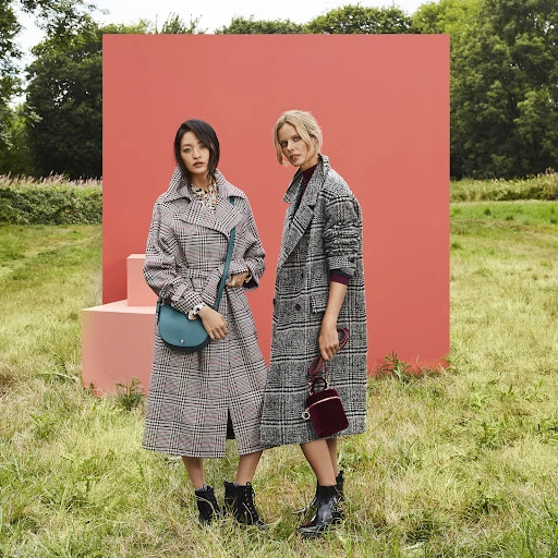 Two women in plaid coats posing in a field in front of a coral-colored backdrop with handbags in a fashion shot.