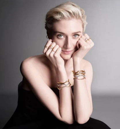 Portrait of a blonde woman wearing multiple gold rings and bracelets, resting her chin on her hands against a grey gradient background.