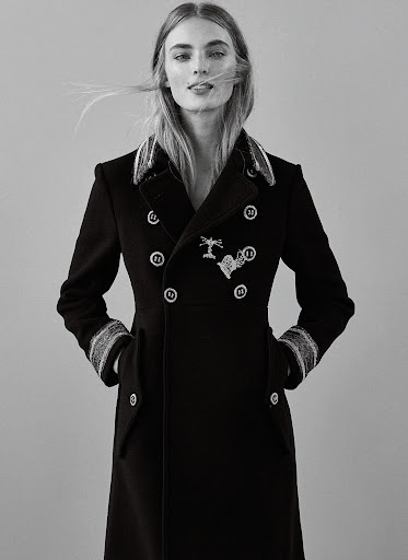 Model wearing a structured double-breasted black coat with decorative buttons against a plain grey background