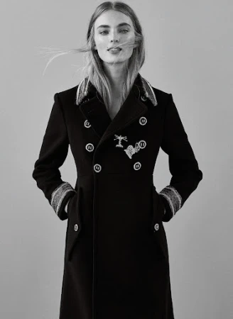 Model wearing a structured double-breasted black coat with decorative buttons against a plain grey background