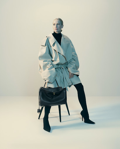 Woman in oversized beige trench coat and black heels seated on stool holding dark leather handbag