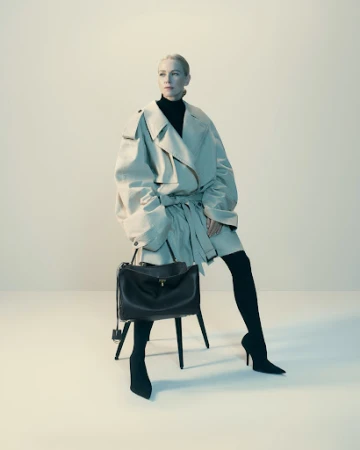 Woman in oversized beige trench coat and black heels seated on stool holding dark leather handbag
