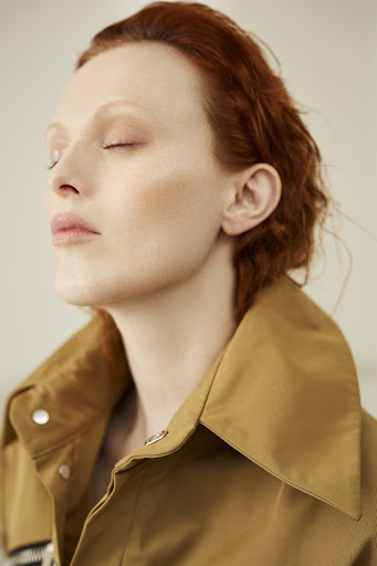 Close-up portrait of a red-haired adult female with eyes closed, wearing a tan collared coat against a neutral backdrop in soft lighting.