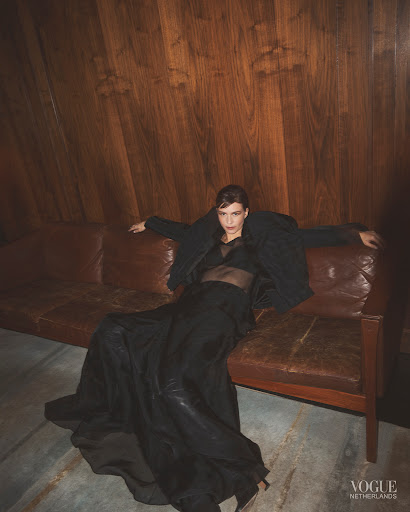 A woman in a sheer top and long black skirt poses on a brown leather sofa in front of a paneled wooden wall, Vogue Netherlands logo is visible.