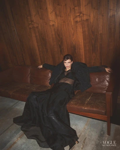 A woman in a sheer top and long black skirt poses on a brown leather sofa in front of a paneled wooden wall, Vogue Netherlands logo is visible.