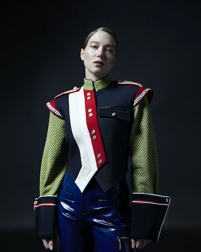 Portrait of a woman wearing a structured military-style jacket with red and white sash details and blue glossy trousers against a dark background.