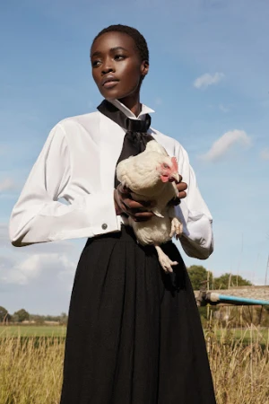 Black woman in a white button-down shirt and black skirt holding a white chicken against a blue sky