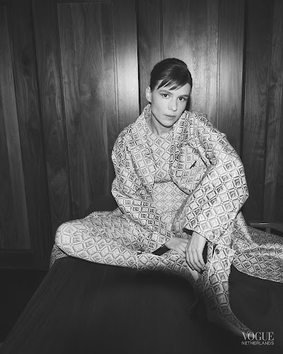 Monochrome portrait of a fashion model in patterned robe and pants sitting against a wood panel wall in a studio environment.