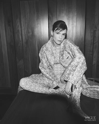 Monochrome portrait of a fashion model in patterned robe and pants sitting against a wood panel wall in a studio environment.