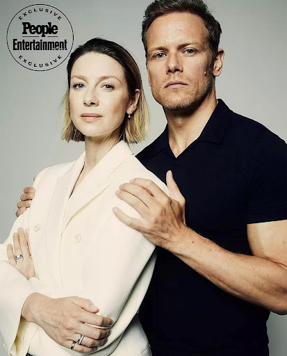 Caitriona Balfe and Sam Heughan pose for a portrait with arms around each other in front of a neutral grey background, shot at eye level.