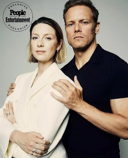 Caitriona Balfe and Sam Heughan pose for a portrait with arms around each other in front of a neutral grey background, shot at eye level.