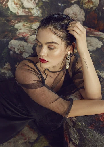 Adult woman with red lipstick poses in sheer black dress with floral patterned background. Close up view, looking off camera.