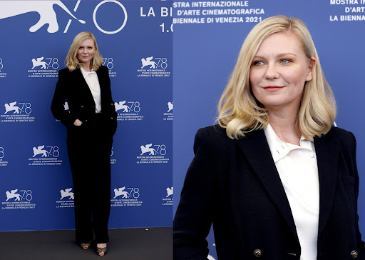 Kirsten Dunst poses on a blue backdrop in a black suit jacket and matching pants with a white top and blonde wavy hair. 