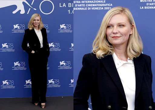 Kirsten Dunst poses on a blue backdrop in a black suit jacket and matching pants with a white top and blonde wavy hair. 