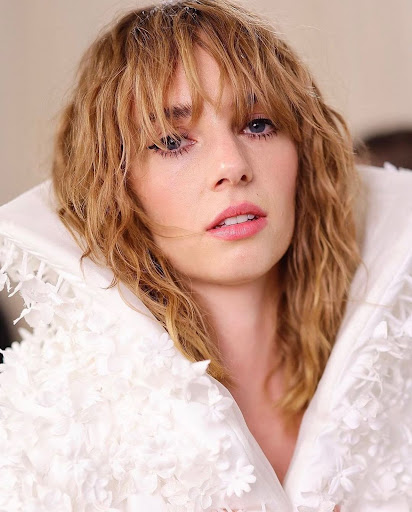 Close-up portrait of a blonde woman with bangs, wearing a white, floral-textured garment, gazing directly at the camera in a soft, diffused light.
