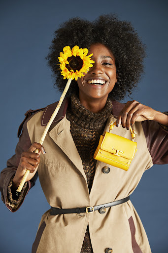 Stylish woman in a trench coat smiles while holding a sunflower and yellow purse against a dark blue studio backdrop.