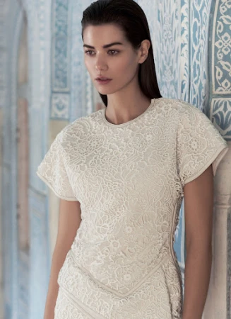 Model with slicked-back dark hair wearing a cream lace dress standing against blue patterned architectural walls