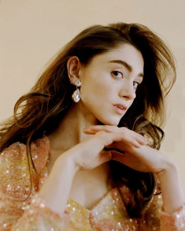 Portrait of a woman wearing teardrop crystal earrings and a sequined top against a plain beige background.