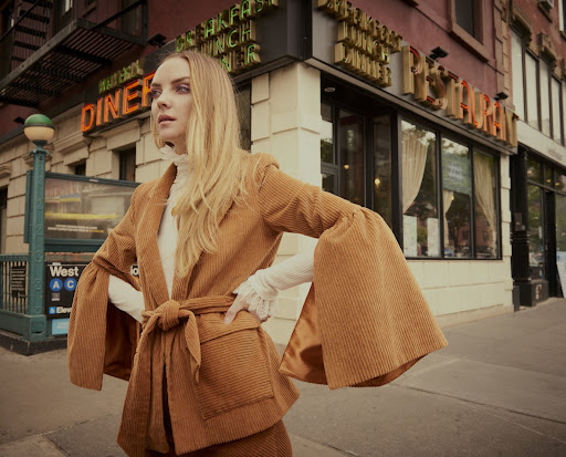 Woman in stylish brown corduroy suit standing on a city street near a diner and subway entrance, posing with hands on hips.