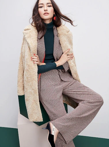 A woman poses in a houndstooth suit and dark green turtleneck, complemented by a beige shearling coat and high heels, against a neutral backdrop.