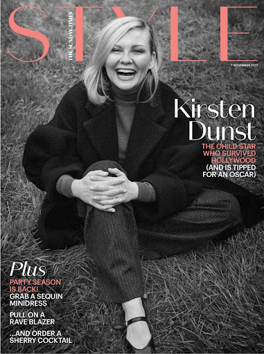 Magazine cover featuring Kirsten Dunst smiling in a dark coat and trousers, seated on the grass with a peach masthead on a black and white photo.