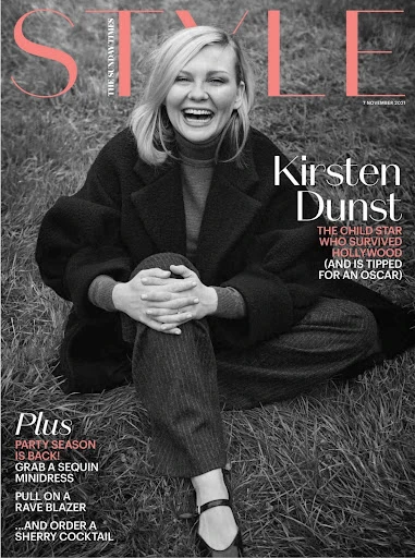 Magazine cover featuring Kirsten Dunst smiling in a dark coat and trousers, seated on the grass with a peach masthead on a black and white photo.