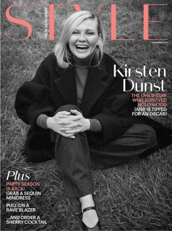 Magazine cover featuring Kirsten Dunst smiling in a dark coat and trousers, seated on the grass with a peach masthead on a black and white photo.