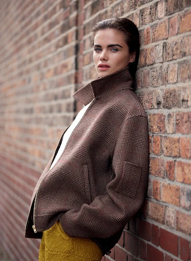 Stylish woman in a brown plaid jacket leans against a brick wall, showcasing a blend of fashion and urban backdrop in natural lighting.