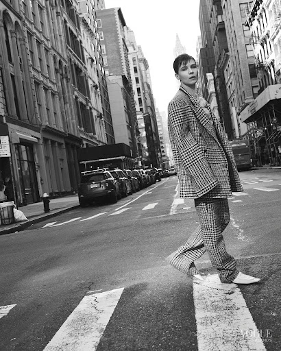 Monochrome outdoor fashion shot: Woman in plaid suit walks on crosswalk in urban setting with tall buildings and parked cars.
