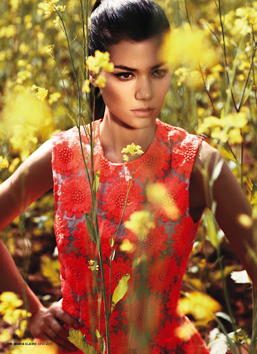 A woman poses in a vibrant coral dress, surrounded by a lush field of yellow flowers under bright lighting. Fashion editorial style.
