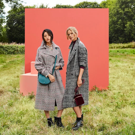 Two female models in plaid coats standing in a grassy field with a coral backdrop, styled by make-up artist Mary Wiles.