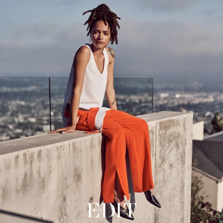 A woman with dreadlocks wearing a white top and vibrant orange trousers seated on a concrete rooftop ledge overlooking a city.