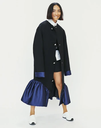 Woman with dark bob haircut wearing black coat with blue satin cuffs and hem, smiling against a white backdrop.