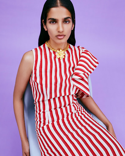 Elegant woman in red and white striped dress poses in front of light purple background, wearing a gold statement necklace.