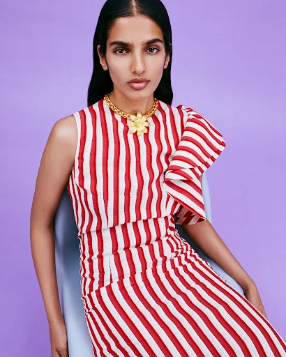 Elegant woman in red and white striped dress poses in front of light purple background, wearing a gold statement necklace.
