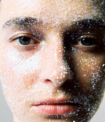 Close-up portrait of a person's face covered in sparkling glitter makeup, emphasizing the eyes and texture of the glitter.