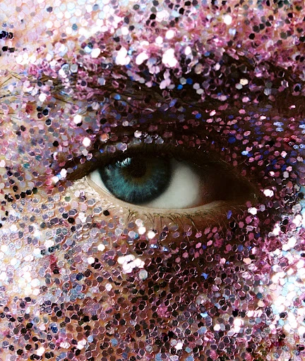 Close-up of a person's face with pink and purple glitter makeup covering the skin and eyebrow, emphasizing the blue-green eye.